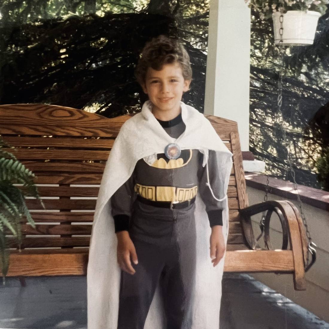A boy in a Batman costume and a white cape standing on a porch