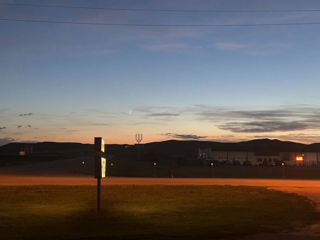 A lit roadside sign under a crescent moon at dusk, plains behind it