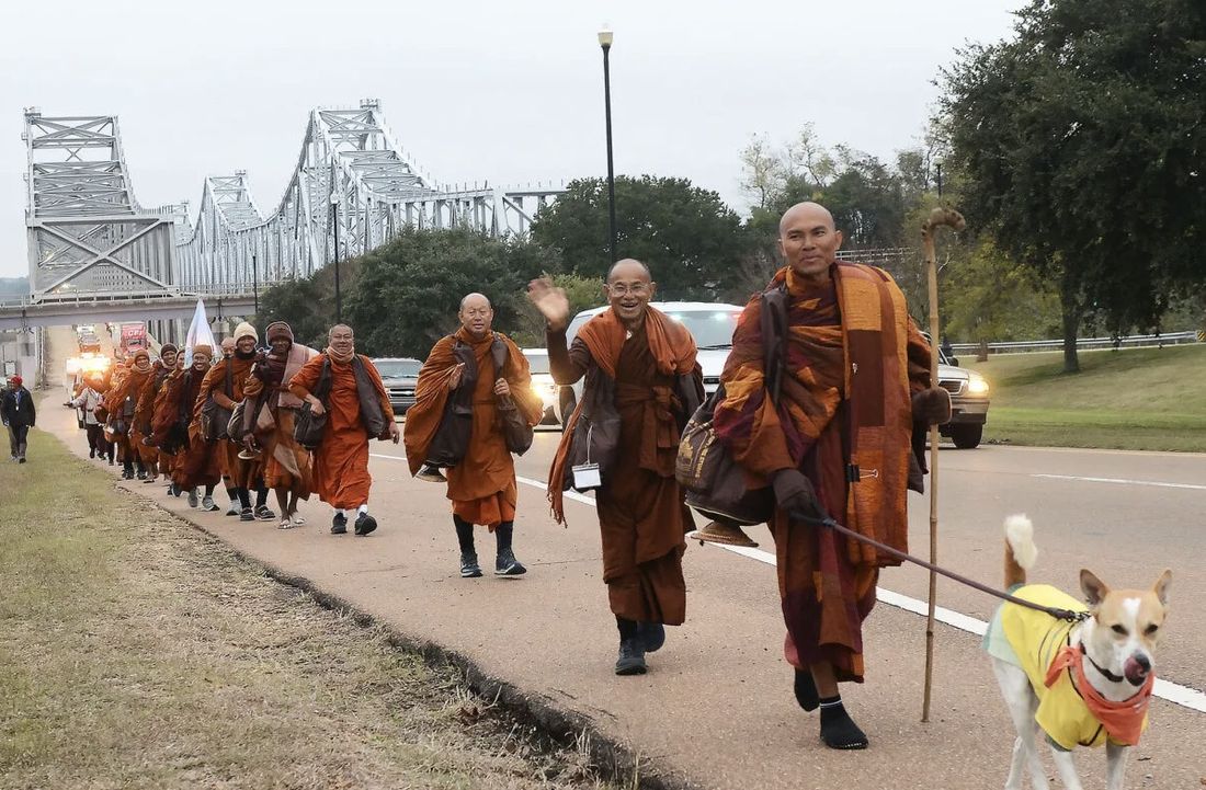 A long line of monks in ochre robes walking a highway shoulder, a dog leading