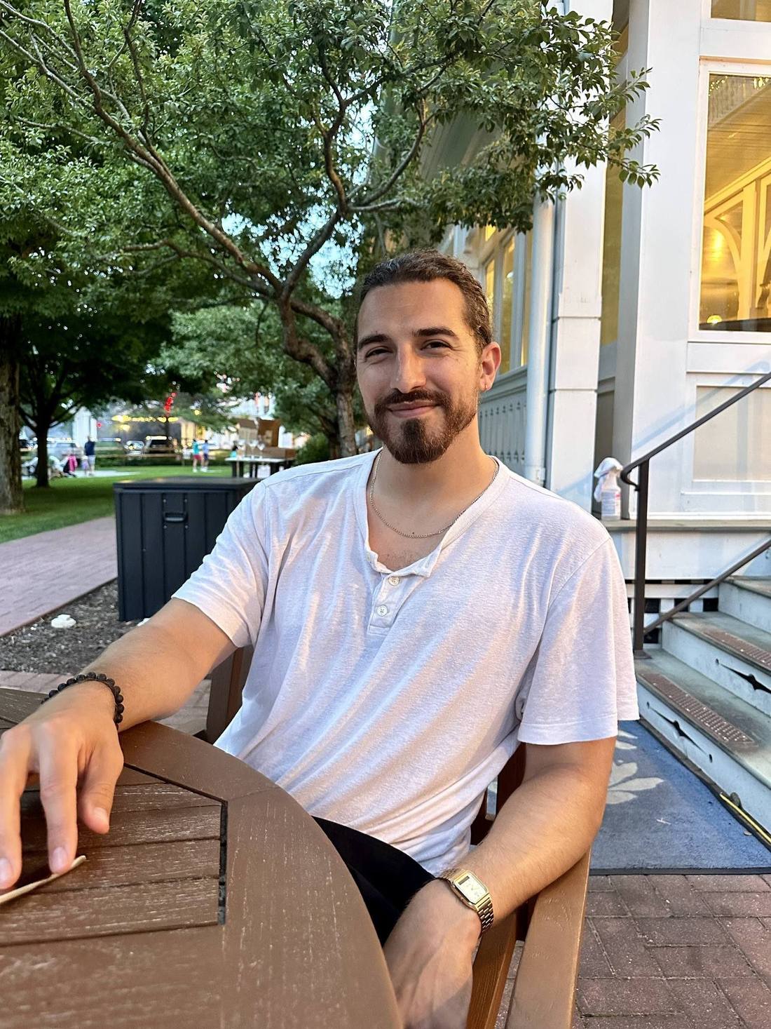 At an outdoor table in a white tee under summer trees