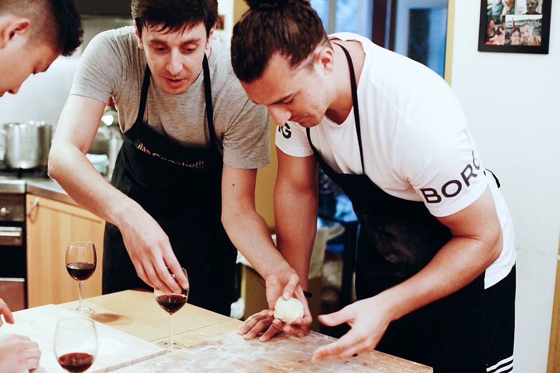 Hands working dough on a floured wooden table, wine glasses beside it