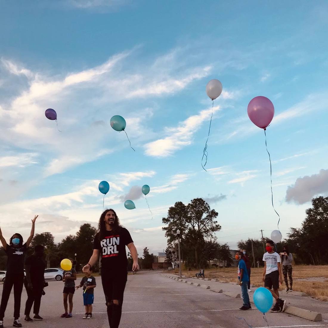 In a parking lot at dusk as a dozen balloons rise, kids watching