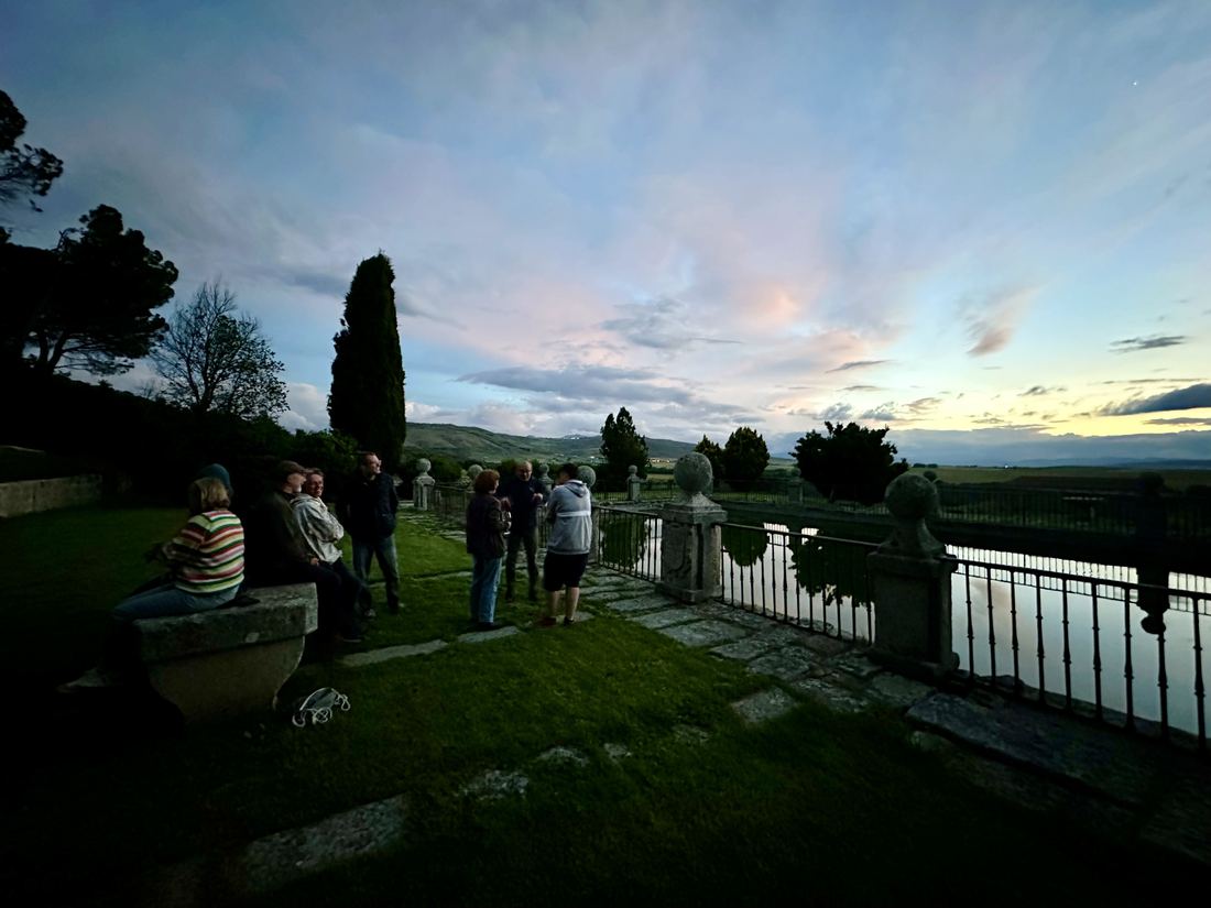 People talking on a terrace above still water at dusk, hills beyond