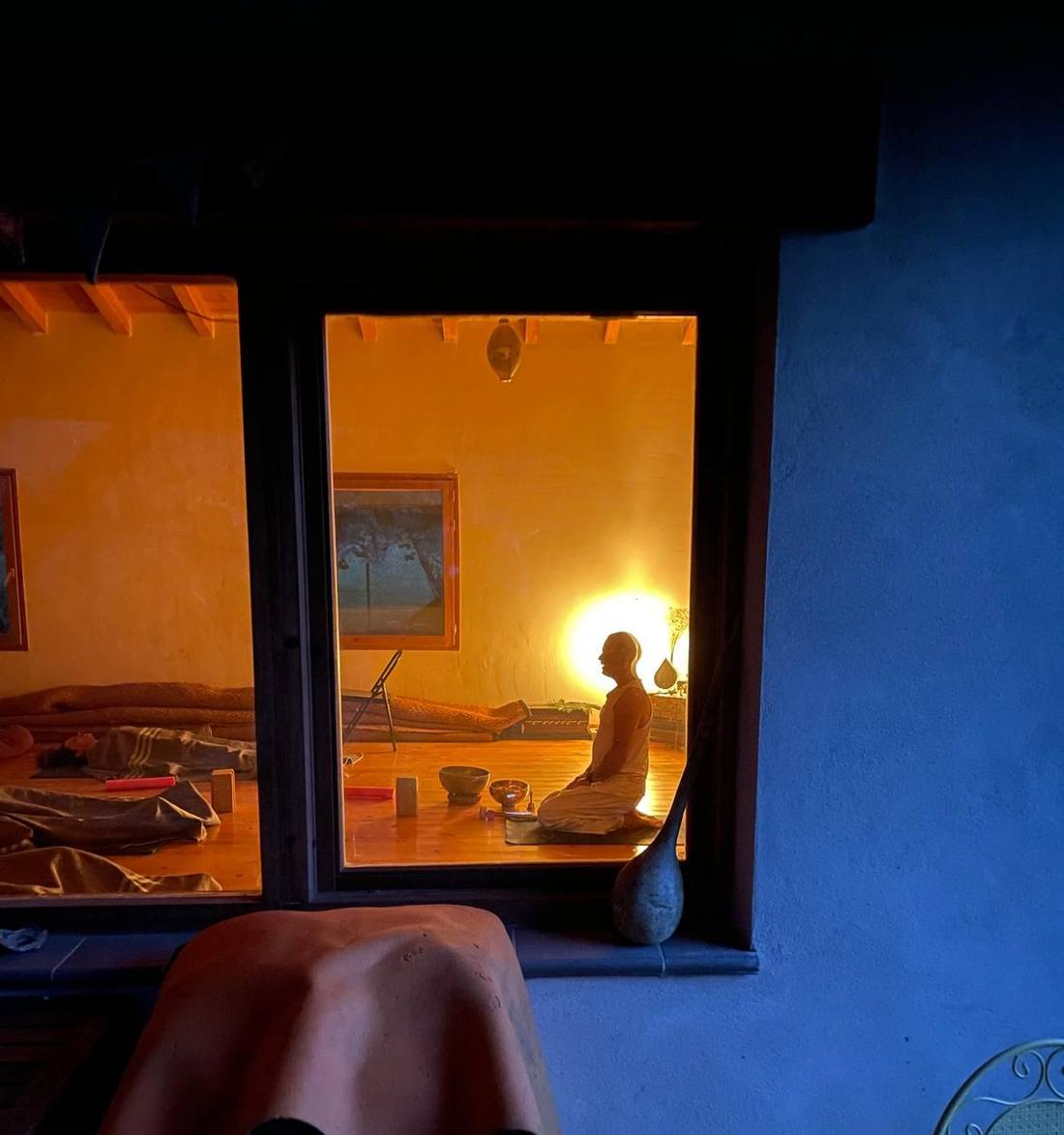 Through a window at night, Roland kneeling in lamplight, blankets on the floor